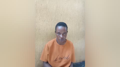 Video of a young Black man with short hair, wearing an orange T-shirt with \"less\" written in black cursive, standing against a textured beige wall. He has a serious expression.