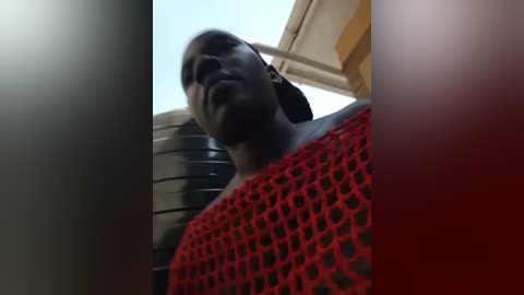 Video of a person in a red, fishnet-patterned top, standing outdoors under a clear sky with a building in the background. The image is low-angle, emphasizing the subject's face and upper body.