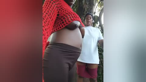 A video of two women in a lush, green outdoor setting. One woman, with fair skin and a large belly, wears a red crochet top and brown pants. The other, with darker skin, is in a white t-shirt and red shorts, standing behind her.