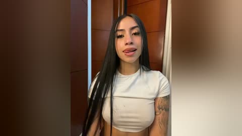 Video of a young Latina woman with long, straight black hair, light skin, and medium-sized breasts, wearing a white crop top. She stands in a doorway with wooden panels, smiling.