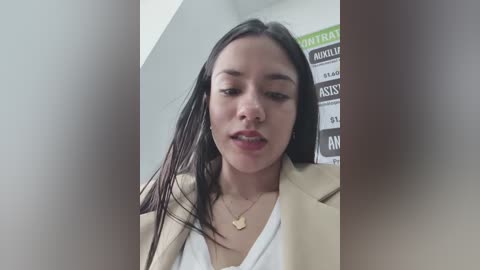 A video of a young Asian woman with long black hair, wearing a beige blazer over a white top, standing in a room with a partially visible green and black sign.