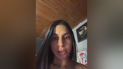 Video of a young woman with long black hair, medium brown skin, and a slightly open mouth, taken indoors with a wooden ceiling and colorful wall art in the background.