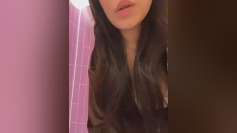 Video of a woman's face and upper chest, with long, wavy dark hair, pink lips, and a black jacket, set against a pink-tiled bathroom wall.