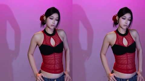 Video of a young Asian woman with medium skin tone, black hair in a bun, wearing a red, sleeveless, mesh top with black accents, and blue jeans, against a pink backdrop.