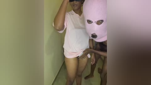 Video of a slim, dark-skinned woman in a light pink t-shirt and no pants, wearing a pink ski mask, standing in a dimly lit, beige-walled corridor.