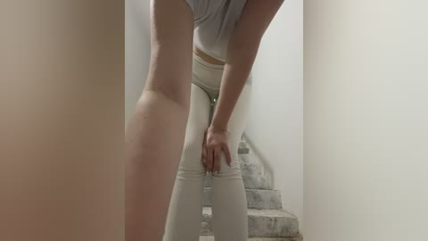 A video shows a woman standing on a staircase, wearing a grey top and tight white pants, leaning against the wall with her hands on her thighs.