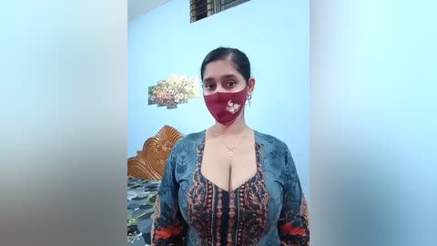 Video of a South Asian woman with dark hair, wearing a red mask, floral dress, and denim jacket, standing in a room with light blue walls, a decorative wall panel, and a wooden headboard.