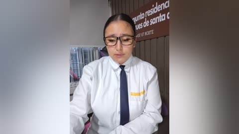 Video of a serious-looking woman with glasses, wearing a white shirt and black tie, standing in front of a bulletin board with Spanish text.