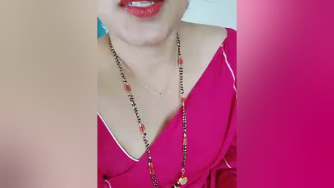 A video of a light-skinned woman with red lipstick, wearing a pink V-neck top and a beaded necklace. The image is partially blurred, with a pink background.