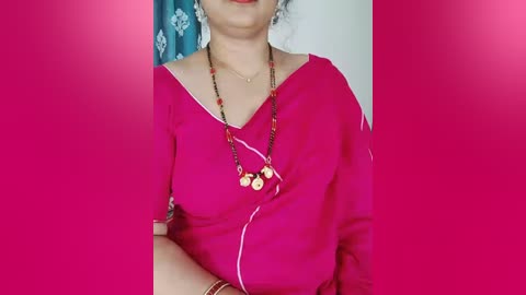 Video of a woman in a vibrant pink saree with a black border, wearing a gold necklace and a red bindi, standing against a magenta background with a teal curtain in the background.