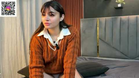 A video of a young woman with long dark hair, wearing an orange sweater and white blouse, sitting on a bed with gray bedding, in a modern room with a dark headboard.