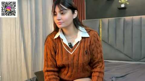 Video of a young Asian woman with straight black hair, wearing a brown sweater over a white collared shirt, sitting on a bed in a modern bedroom with gray walls and curtains.