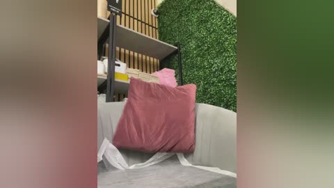 A video of a messy room with a pink pillow on the floor, surrounded by green artificial plants, cardboard boxes, and white plastic sheets, creating a cluttered, disorganized environment.
