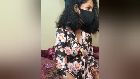 Video of a South Asian woman with medium-dark skin, wearing a floral-printed blouse, black face mask, and headscarf, sitting on a bed with pink and red floral bedspread.