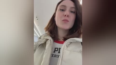 A video of a young woman with shoulder-length brown hair, fair skin, and a neutral expression, wearing a white jacket and a red-and-white shirt. The image is blurry with a focus on her face, taken from a low angle.