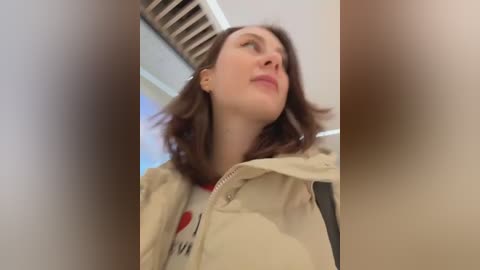 Video of a woman with shoulder-length brown hair, wearing a beige jacket, standing in a modern, brightly lit room with a ceiling fan.
