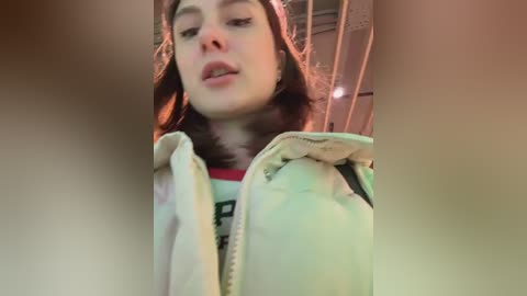 Video of a young woman with fair skin, brown hair, and closed eyes, wearing a pastel-colored jacket, standing indoors near a staircase.