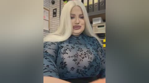 Video of a curvy, fair-skinned woman with platinum blonde hair, wearing a sheer blue floral top revealing her ample breasts, in an office setting with filing cabinets and paperwork.