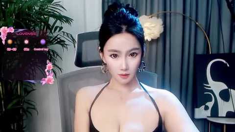 Video of a young East Asian woman with fair skin, black hair in a bun, wearing a black halter top, sitting in a modern room with a gray chair, plants, and abstract art.