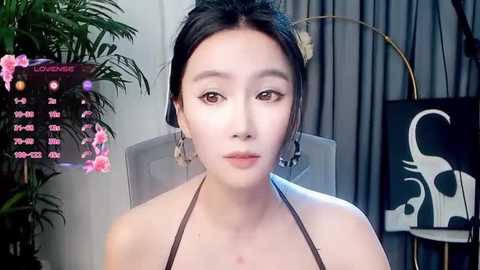 Video of a young East Asian woman with fair skin, dark hair in a bun, wearing a brown halter top, seated in a modern room with a calendar, greenery, and abstract art.