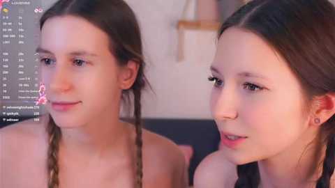 Video of two young women with fair skin and long brown hair, one braided, sitting close together, smiling. Background includes a blurred, neutral-colored room and a smartphone screen displaying social media notifications.