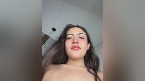 Video of a young woman with long, dark hair, wearing a light-colored top, and a thin necklace. She has fair skin, full lips, and is indoors with a ceiling fan visible in the background.