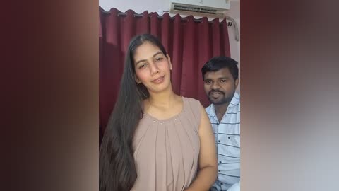 Video of a smiling South Asian woman with long black hair, wearing a beige sleeveless top, standing beside a man with short black hair in a white striped shirt, both in front of a red curtain backdrop.