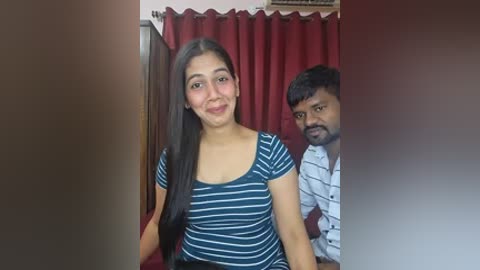 Video of a smiling South Asian woman with long dark hair, wearing a blue striped top, standing next to a man with a mustache in a room with red curtains.