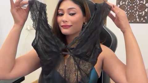 Video of a Latina woman with long brown hair, light skin tone, wearing black lace lingerie, smiling, holding the garment, in a modern office setting with a black leather chair and white wall.