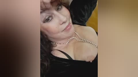 A video shows a middle-aged woman with fair skin, curly dark hair, and large breasts partially exposed, wearing a black velvet dress and pearl necklace. The image is slightly blurred.