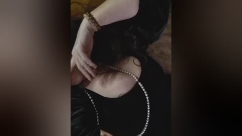 A video of a woman's upper body, showing a glimpse of her black dress and pearl necklace, with a bracelet on her wrist, her hand resting on her chest. The background is blurred.