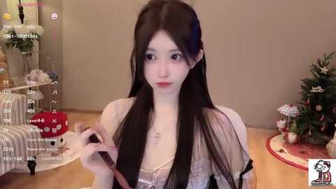 Video of an Asian woman with long black hair, pale skin, wearing a sheer white dress, sitting on a wooden floor, playing with a red toy in a room with a Christmas tree.