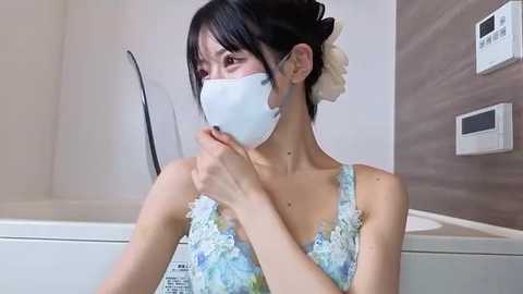 Video of a young Asian woman in a pastel blue floral dress with a floral face mask, seated in a modern bathroom with a white tub and gray wall.