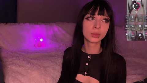Video of a pale woman with long black hair and bangs, wearing a black top, sitting on a white faux fur rug, in a dimly lit room with purple lighting and a glowing Apple logo.