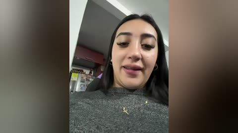 Video of a Latina woman with long black hair and light skin, wearing a gray sweater, standing in a dimly lit room with shelves and a red object in the background.