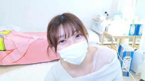 Video of a young East Asian woman with brown hair, wearing a white face mask, white top, and sitting in a bright, minimalist room with a pink bedspread, wooden table, and a blue and white bag.