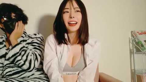 Video of a smiling East Asian woman with straight black hair, wearing a Calvin Klein bra and a white shirt, seated next to a person in a zebra-patterned blanket.
