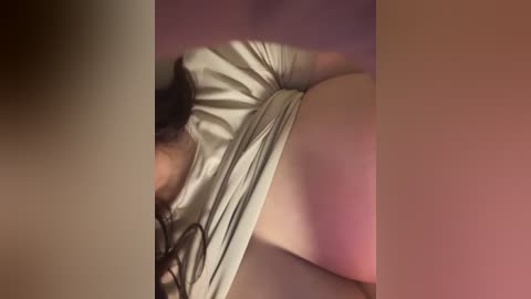 Video of a woman in a beige top, lying on her side, her dark hair partially obscuring her face, with soft, warm lighting creating a sensual ambiance.