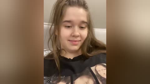 Video of a young, fair-skinned girl with long, brown hair, wearing a black shirt with a cartoon character, smiling. Background shows a beige wall and a white bathtub.