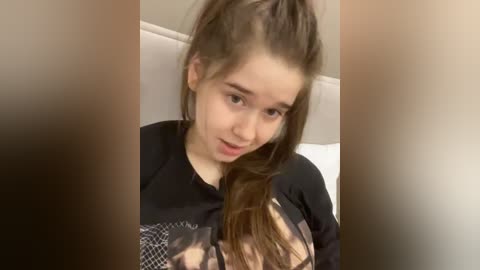 A video of a young Caucasian girl with long brown hair in a high ponytail, wearing a black long-sleeved shirt, lying on a white bed with a beige headboard.