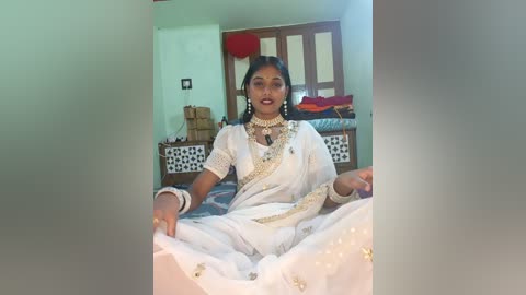 Video of a woman in traditional Indian attire, white saree with gold embroidery, seated cross-legged on a bed, wearing gold jewelry, in a modestly furnished bedroom with green walls and wooden furniture.