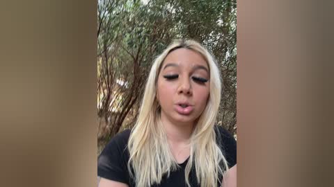 Video of a young, fair-skinned, blonde woman with long hair, wearing a black top, pouting outdoors amidst greenery.