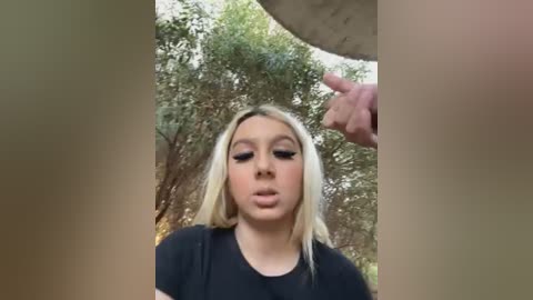 Video of a young blonde woman with makeup, wearing a black top, looking at a man's hand in a forest background.