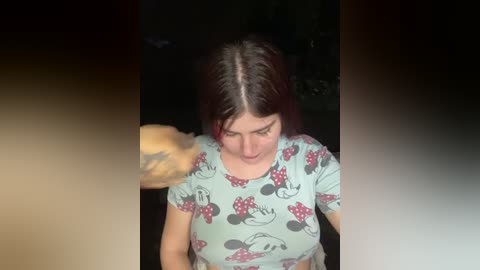 Video of a young woman with straight, dark hair, wearing a light blue t-shirt with Mickey Mouse and grapes pattern. Her expression is neutral. Background is dark and blurry.