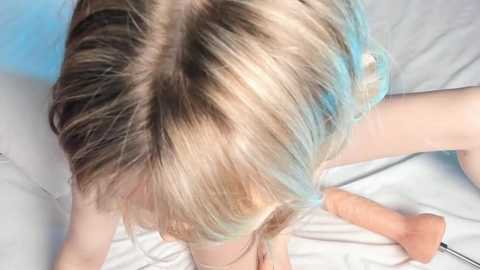 A video shows a blonde woman with a blue streak in her hair, wearing a white dress, lying on a bed, with a flesh-colored dildo beside her.