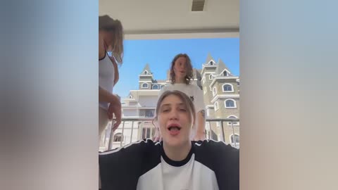 Video of a young girl with short brown hair, wearing a black and white sports shirt, being filmed by a blonde woman on a balcony overlooking a Victorian-style mansion.
