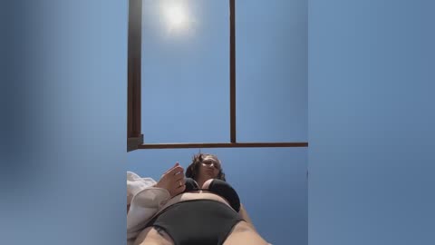 Video of a woman in a black bra and grey panties lying on a bed, gazing upward at a blue sky through a window.