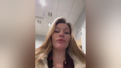 Video of a young woman with long, straight blonde hair, wearing a beige jacket and a dark shirt, looking slightly off-center. The background shows a dimly lit room with a white ceiling and recessed lights.