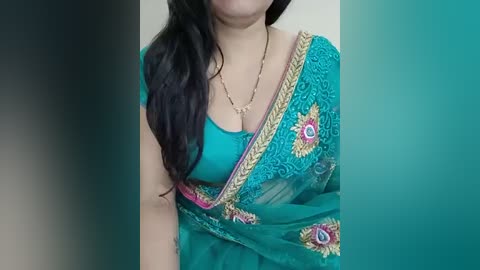 Video of a woman with long black hair, wearing a turquoise sari with intricate pink and gold embroidery, a teal blouse, and a gold necklace. Background is blurred.