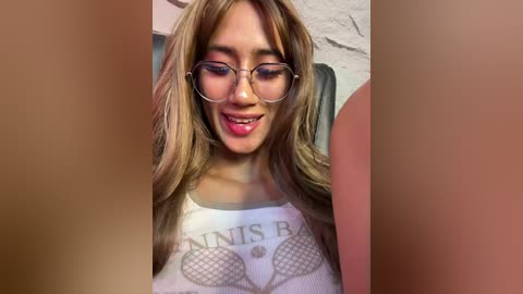 Video of a young woman with light skin, long brown hair, wearing round glasses, and a white \"Bee's Knees\" tank top. She is smiling.